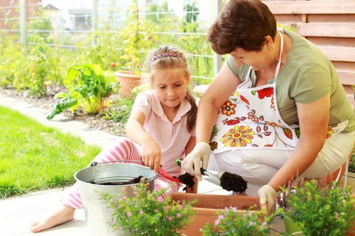 A woman and a young girl are gardening together outdoors in a neatly maintained backyard. The woman is kneeling beside the girl, both wearing gardening gloves, and they are planting or tending to plants in a rectangular wooden garden bed. The garden bed is filled with dark soil, and nearby there are potted plants and flowering shrubs, adding colour and variety to the garden. In the background, a lush green lawn extends to a wire fence with additional garden plants and trees, indicating a well-kept outdoor space typical of residential gardens in Willesden. The scene is set on a bright day with natural sunlight illuminating the area, showcasing an inviting outdoor environment suitable for family gardening activities, which complements the gardening services offered by Landscapers Willesden in the local area.