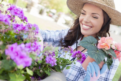 A woman with long brown hair, wearing a wide-brimmed straw hat, a blue and white checkered shirt, and light blue gardening gloves, is smiling as she tends to a colorful flower bed in a well-maintained garden. The flower bed contains a variety of vibrant blooming flowers in shades of purple, pink, and peach, with lush green foliage surrounding them. In the background, there is a neatly trimmed lawn with the edge of a paved patio or pathway visible, along with some trees and outdoor furniture, suggesting a landscaped outdoor space typical of a suburban garden. The scene is set in natural daylight, indicating a clear and pleasant weather condition, with soft sunlight illuminating the garden. This garden layout includes a prominent flower bed with rich soil borders, complemented by a tidy lawn area, reflecting professional gardening and landscaping care, such as those offered by Landscapers Willesden, with a natural and inviting outdoor environment perfect for leisure and outdoor activities.