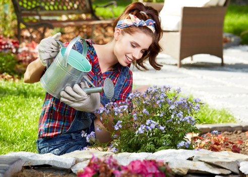 A young woman with a cheerful expression is gardening outdoors in a well-maintained garden, kneeling next to a flower bed filled with blooming purple and pink flowers. She is wearing a plaid shirt, denim jeans, gloves, and a headband, and is watering the plants with a silver watering can. Behind her, there are lush green trees, a paved pathway, and garden furniture, indicating a cared-for outdoor space typical of residential gardens in Willesden. The scene is brightly lit with natural daylight, suggesting a spring or summer day, and highlights the garden's mix of vibrant flowering plants, neatly edged flower beds, and surrounding greenery, reflecting professional gardening and landscaping efforts. This setting illustrates the types of outdoor maintenance and gardening services offered by Landscapers Willesden for enhancing residential outdoor spaces in London.