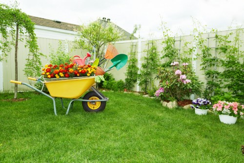 A well-maintained garden in Willesden with a lush, green lawn occupying the majority of the space, bordered by a white wooden fence covered in climbing plants and foliage. In the foreground, a yellow wheelbarrow filled with vibrant flowers and garden tools such as a watering can and small spade is positioned on the grass. To the right, there are several flower beds with blooming plants in shades of pink, purple, and white, arranged neatly along the border of the yard. A small shrub or young tree stands near the fence, providing vertical interest, while other foliage adds texture to the scene. The overall environment is bright and natural, suggesting a mild weather day suitable for gardening activities. This image illustrates a typical suburban backyard in Willesden, suitable for landscaping and flower bed creation services by Landscapers Willesden, highlighting the readiness for outdoor enhancements and garden improvement projects.