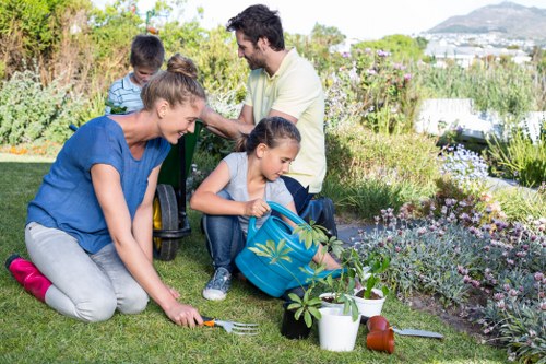 A family outdoors in a landscaped garden with a well-maintained lawn, flower beds filled with blooming plants, and a variety of shrubs and small trees. The scene shows a woman kneeling on the grass, planting or tending to a small plant, while a young girl assists by holding a gardening trowel and a blue bag. Nearby, garden tools like a watering can and a gardening fork are visible on the grass, suggesting active gardening work. In the background, a man and a boy sit on the grass, watching and enjoying the outdoor space, which features a mix of flowering plants and greenery. The garden appears to be part of a residential property, with a scenic view of distant hills and a sky with natural daylight, ideal for outdoor family activities and garden maintenance. The overall environment indicates a well-kept, inviting outdoor area maintained by professional landscapers such as Landscapers Willesden, suitable for gardening, lawn care, and outdoor landscaping services in the local area near Willesden, London.