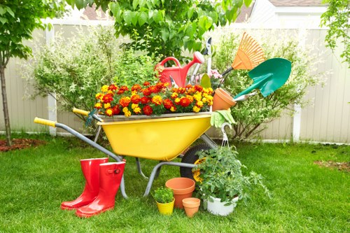 A garden scene featuring a vibrant yellow wheelbarrow filled with red and orange flowering plants positioned on a well-maintained green lawn. Nearby, there are red rubber boots resting on the grass, and several terracotta and plastic plant pots, with a potted shrub nearby. In the background, leafy green trees and shrubs provide a lush backdrop, with a white fence enclosing the garden area. Gardening tools, including a watering can, a rake, and a bright green trowel and shovel, are also visible, leaning against the wheelbarrow. The overall scene captures a tidy and inviting outdoor space suitable for gardening services, with natural daylight and a clear sky, illustrating a typical residential garden at a time of active plant care. Landscapers Willesden service this area, supporting garden maintenance and landscaping needs with attention to detail and natural aesthetics.