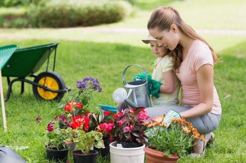 The image depicts a woman and a young girl working together in a well-maintained garden, planting flowers and plants in various pots and containers on a lush, green lawn. In the foreground, there are colourful flowers including reds, purples, and oranges, arranged neatly alongside gardening tools and small watering cans, indicating active outdoor care. To the left, a green garden wheelbarrow with yellow wheels is positioned on the grass, and in the background, a row of shrubs or hedges lines the garden boundary, providing natural structure and privacy. The outdoor environment appears bright and breezy with natural daylight, suggesting a mild or sunny day suitable for gardening. The scene reflects a typical residential garden setup in Willesden, showcasing landscaping and lawn care activities that Lawncare services like those from Landscapers Willesden might support. The combination of flower beds, lawn space, and garden tools highlights the time and effort involved in outdoor maintenance and planting tailored to local garden styles and conditions in the area.