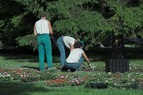 The image depicts three individuals working together in a well-maintained garden, surrounded by lush green trees and a neatly edged flower bed with colorful blooming flowers such as pink, red, and white. One person stands upright holding garden tools, while another is crouched down tending to the soil or plants, and the third is kneeling nearby also engaged in gardening tasks. The garden features a dense hedge or trees in the background, providing shading and privacy, with a lawn area covered in evenly cut, vibrant green grass. To the right, a black plastic garden waste compost bin is visible, indicating sustainable yard management practices. The overall scene highlights active outdoor maintenance and planting efforts in a tidy, urban garden environment, typical of residential outdoor spaces in Willesden, which could benefit from local gardening services focused on lawn and plant care, composting, and sustainable landscaping.