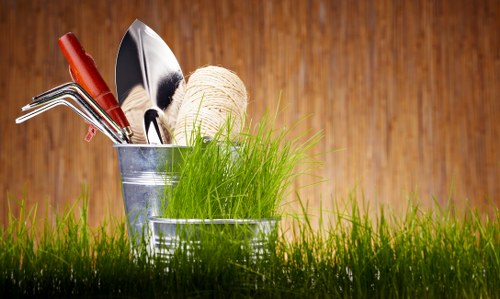 The image depicts a close-up view of a small gardening tool set placed on a lush, green lawn in a garden environment. The tools, including a hand rake, trowel, and a small fork, are neatly arranged inside a metallic container, with a spool of natural twine and fresh, green grass growing out from the container. The lawn features densely packed, healthy grass blades with vibrant natural tones, contrasting with the warm, polished wooden surface in the background. The outdoor setting appears well-maintained, suitable for planting and gardening activities, and the lighting suggests a partly sunny day, highlighting the natural colors and textures of the garden elements. This scene reflects the tools and environment typical of professional gardening services provided by companies like Landscapers Willesden, focusing on outdoor maintenance and sustainable garden care in the area around Willesden, NW London.
