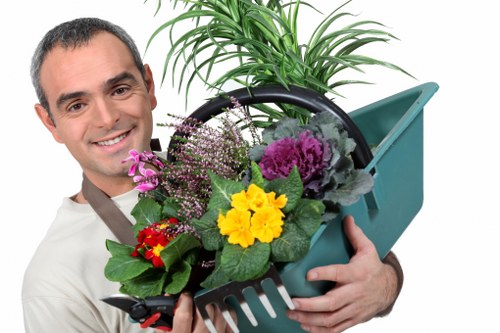 A smiling male gardener holding a green plastic garden fork filled with a variety of vibrant flowering plants such as yellow, purple, red, and pink blooms along with lush green leaves. Behind him, there is a large potted plant with elongated green foliage. The background is plain white, emphasizing the gardening tools and plants. The image reflects outdoor gardening activities typical of landscaping services in the Willesden area, with a focus on plant care and garden maintenance. The professional presentation highlights the company's expertise in garden planting and upkeep in a suburban or urban outdoor space, aligning with landscaping and lawn care services offered by Landscapers Willesden.