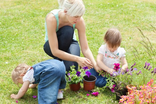 A woman with short blonde hair, dressed in a light blue sleeveless top and dark jeans, is kneeling on a well-maintained green lawn, engaged in gardening activities with two young children. One child, with short light brown hair and wearing a white shirt with pink floral patterns, is sitting on the grass, closely inspecting or tending to a small flower bed with vibrant pink flowers. The other child, with light blonde hair and wearing a blue dress, is also on the grass, reaching towards the soil or plants. The scene includes various gardening tools and pots filled with flowering plants, situated in a landscaped outdoor area that appears to be part of a front or back garden in Willesden, London. The grass is lush and evenly cut, with a backdrop of additional greenery, suggesting a family-oriented, well-maintained outdoor space suitable for gardening and outdoor activities, reflecting professional gardening and landscaping upkeep by Landscapers Willesden.