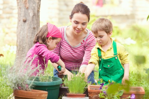 A woman with dark hair tied back, dressed in a pink and white striped top, is outdoors in a garden with two young children, one girl in a pink headscarf and a purple jacket, and one boy in a yellow shirt and green overalls. They are engaged in gardening activities around a table with various potted plants, including lavender, herbs, and flowering plants, set against a background of trees, a hedge, and a garden fence. The scene is illuminated by natural daylight, with lush green foliage and flowering plants visible, suggesting a well-maintained outdoor space suitable for gardening and landscaping activities in Willesden, London. The garden layout includes a mix of flower beds and outdoor plant containers combined with natural grass areas and paved pathways, indicating a thoughtfully designed landscaped yard with opportunities for sustainable outdoor maintenance.