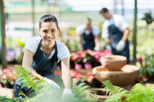 A smiling woman wearing a navy apron and gardening gloves is kneeling in a lush greenhouse filled with vibrant flowering plants and green foliage. She is leaning forward slightly, with her hands resting on a bed of ferns and colorful flowers, clearly engaged in gardening work. Behind her, three other garden workers are visible, dressed in similar attire, tending to potted plants and flower displays, indicating a collaborative gardening effort at a professional landscaping service. The greenhouse features a bright, naturally lit environment with a glass roof and walls, allowing sunlight to illuminate the lush greenery, creating a natural and inviting outdoor atmosphere. The scene showcases a well-maintained garden space with a variety of plant textures, including leafy ferns, flowering plants with pink and red blooms, and neatly arranged flower beds. The background hints at a structured layout, typical of professional horticultural work in a commercial or community garden setting, supported by the presence of gardening tools and organized plant beds, reflecting the expertise of landscapers such as Landscapers Willesden serving the local area, possibly near NW10 postcode.